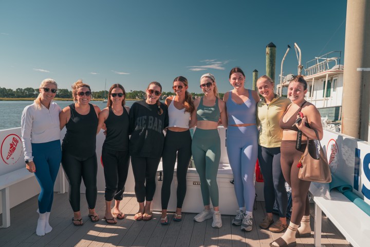 Group of nine women smiling on a boat deck with a clear blue sky.