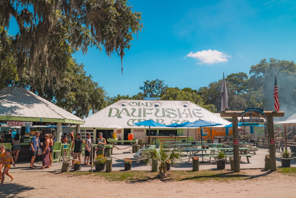 Outdoor market scene at Old Daufuskie Crab Co with people, trees, and blue sky.