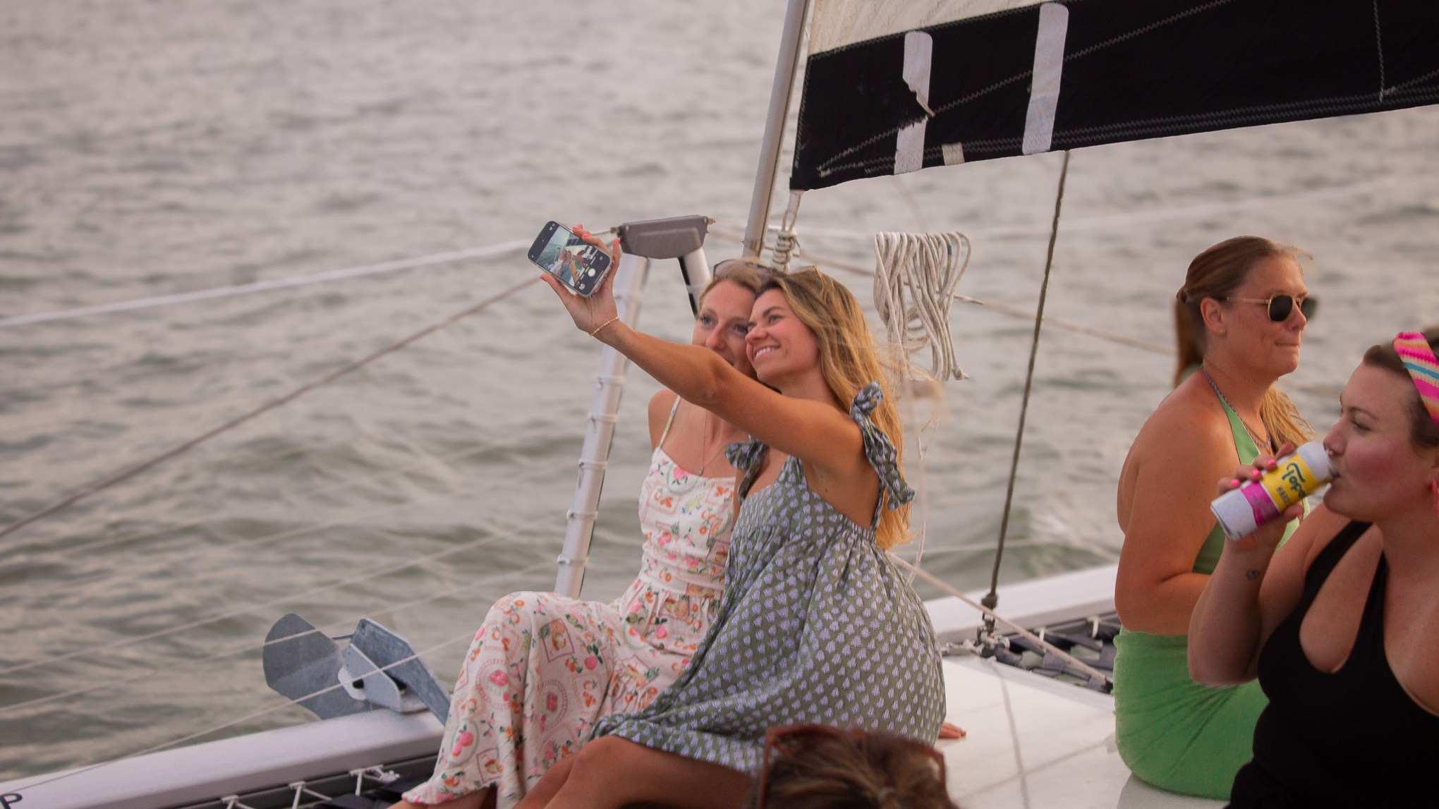 People on a sailboat, with two taking a selfie and one drinking from a can, in front of the ocean.