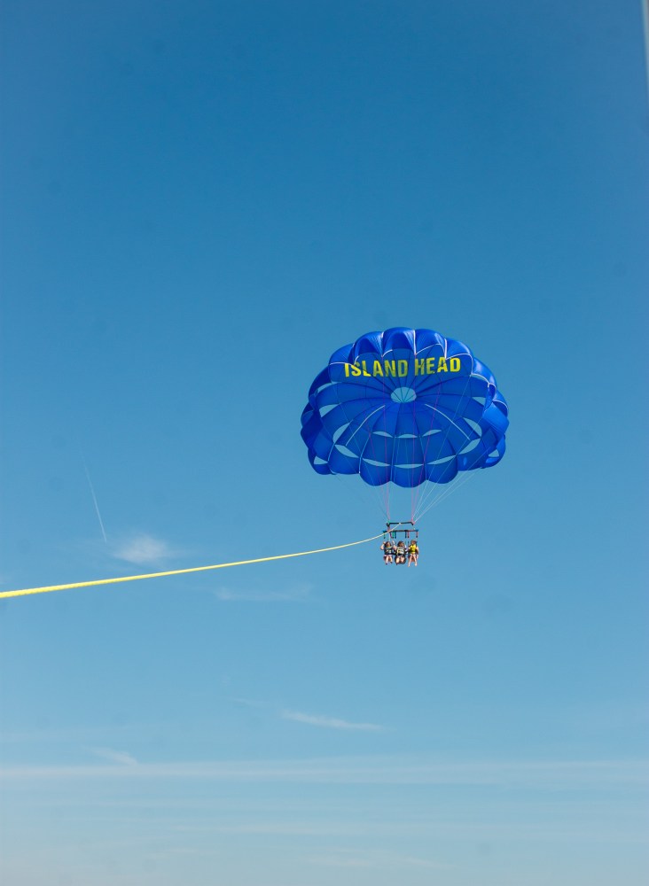 People parasailing with a blue parachute labeled 'ISLAND HEAD' against a clear blue sky.