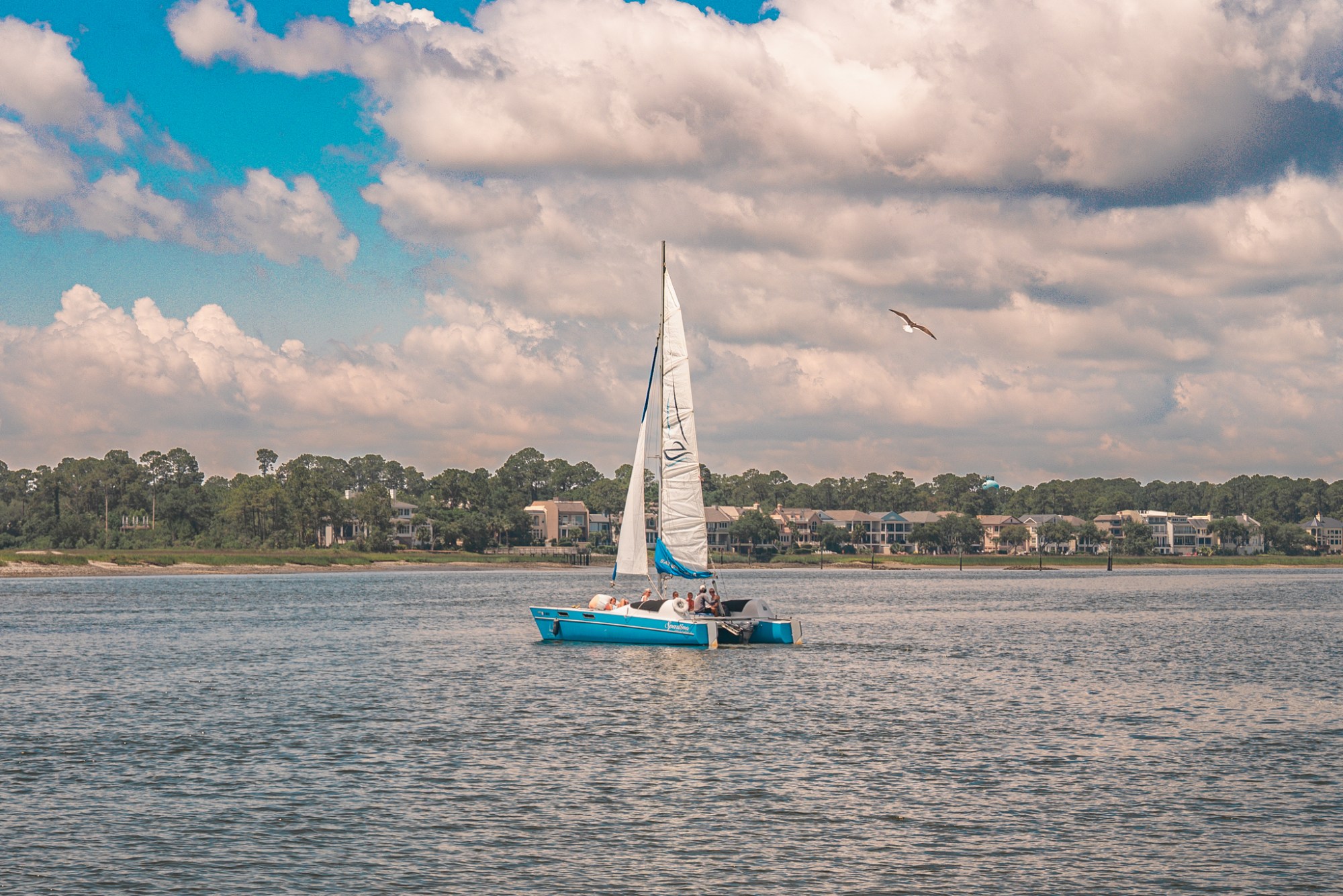 Blue sailboat with people on a lake, distant homes, and a cloudy sky.