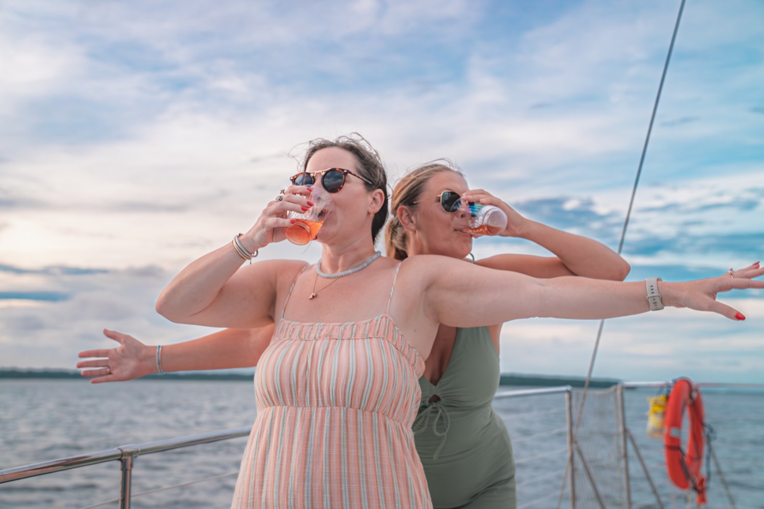 Two women in sunglasses drinking on a boat deck with one striking a pose.