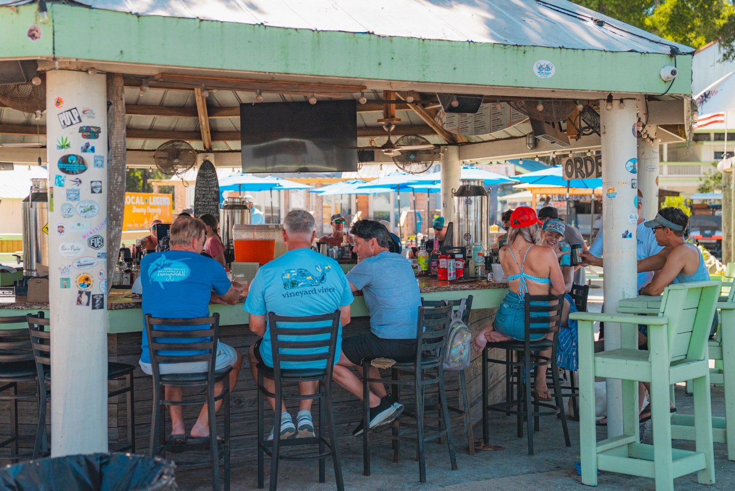 People sitting at an outdoor bar with umbrellas, wearing casual summer clothing.