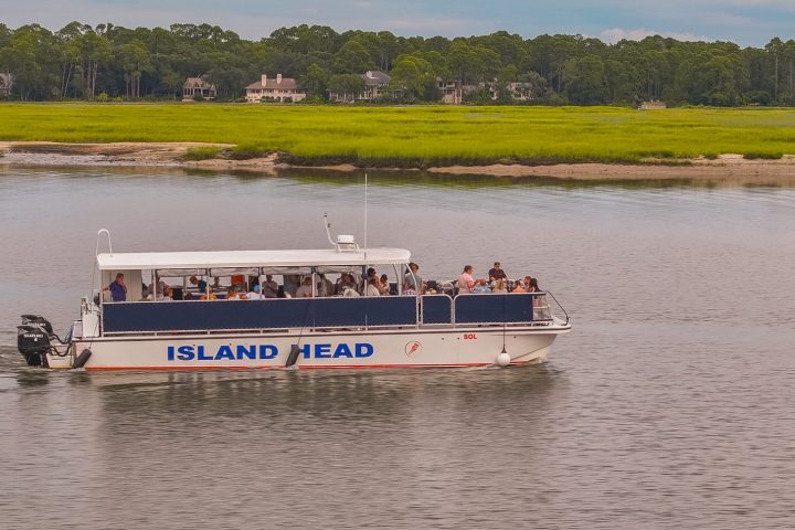 Tour boat named Island Head cruising on a river with a grassy shoreline and trees in the background.