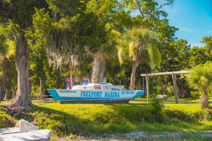 Boat named 'Freeport Marina' displayed on grass, surrounded by trees and swing set.