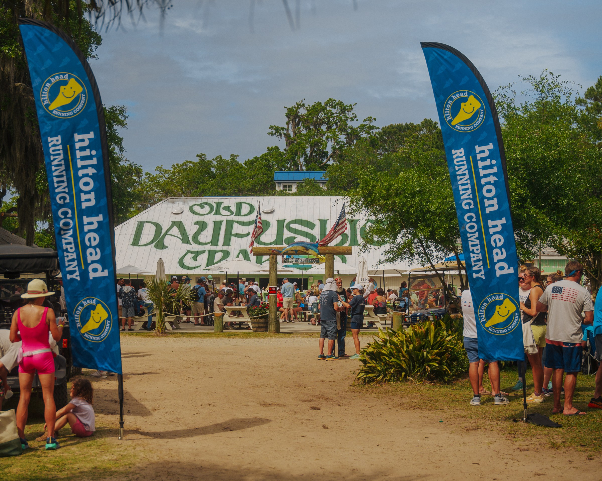 Outdoor event with people, blue banners, and a building labeled 'Old Daufuskie Crab'.