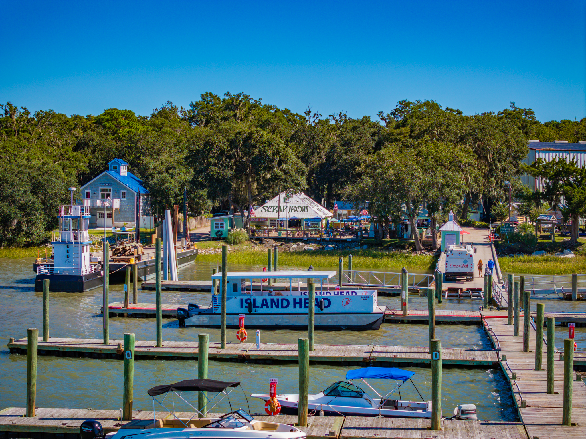 Boats docked at a marina with trees and a building in the background under a clear blue sky.